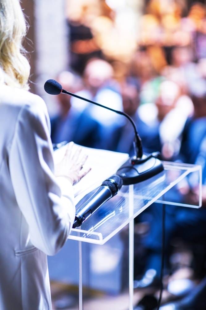 Female,Public,Speaker,At,Business,Conference,,Corporate,Presentation,,Workshop,,Coaching Business professional leading a team meeting in a modern office, presenting a strategic marketing plan to colleagues—highlighting collaboration, campaign planning, and influencer program development.